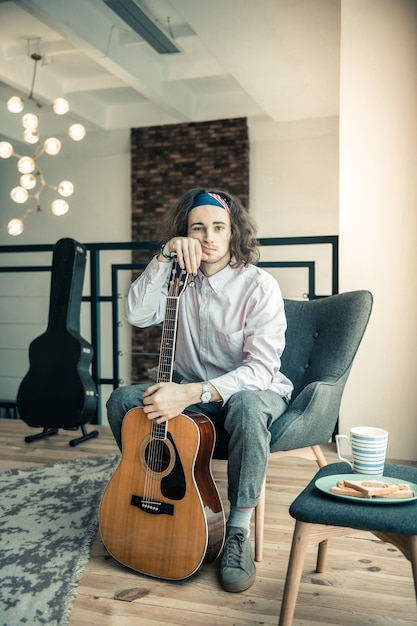 Emotional dark-haired guy. Tired upset guy with colorful bandana leaning on his guitar while sitting in armchair