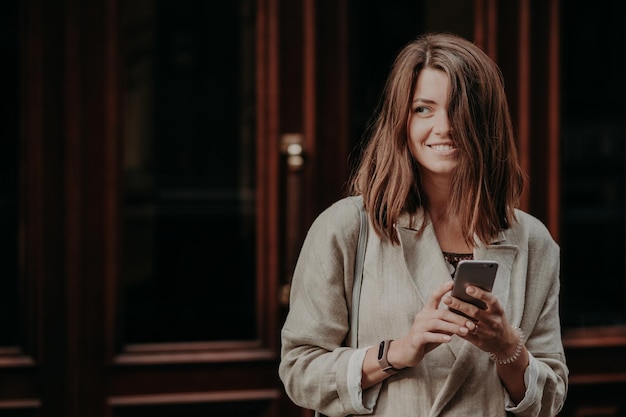 Emotional delighted happy brunette young woman with dark hair, uses mobile phone for texting messages, dressed in elegant raincoat, stands near door, cant imagine life without modern technoliges