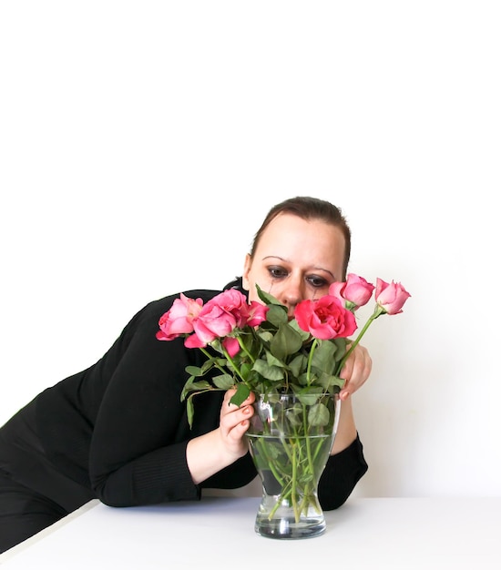 Emotional female portrait on light background of the crying brunette woman in black dress with the bouquet of rose flowers and face with smudged makeup and mascara flowing down her cheeks