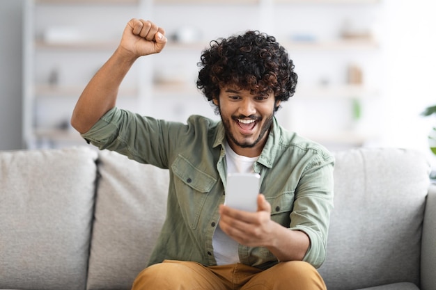 Emotional indian guy holding cell phone raising hand up