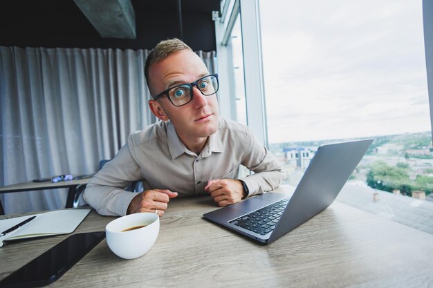 Emotional man looking at the camera at the workplace a man in glasses sitting at a desktop with a computer a student is looking for inspiration for new ideas in an office cafe