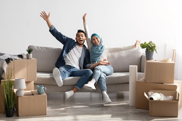 Emotional muslim family celebrating moving to their new house, sitting on new couch among paper boxes with stuff and having break while unpacking belongings, raising hands up and smiling, copy space