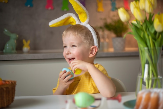 Emotional portrait of a cheerful little boy wearing bunny ears on Easter day who laughs merrily plays with colorful Easter eggs sitting at a table in the kitchen