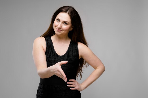 Emotional pretty friendly positive plus size model standing in studio offering handshake on gray background