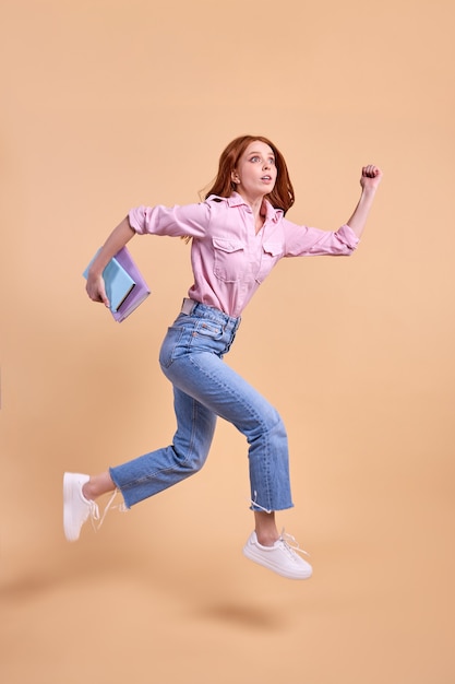 Emotional redhead female student rushing forwards with book in hands, hurry to university, wearing casual outfit, running, 1 September exam, isolated in studio, full-length portrait