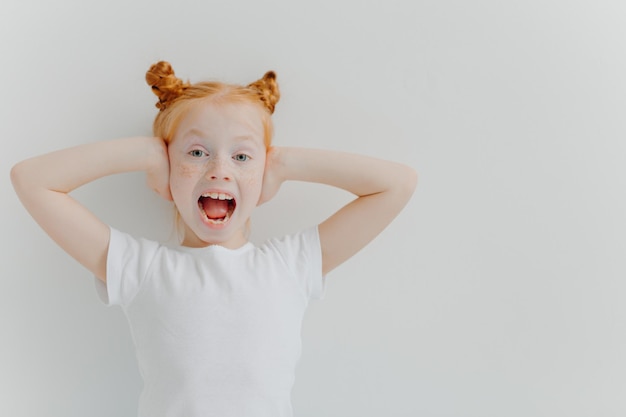 Emotional redhead girl keeps mouth widely opened covers ears excited by noisy atmosphere wears white casual t shirt reacts emotionally on something has fun isolated over white background