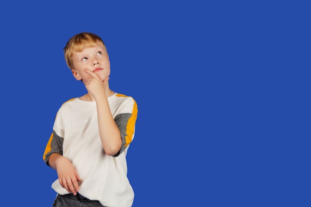 Emotional teenager thinks and holds his hand to his chin in thought, isolated against blue Royal background. Portrait of Caucasian boy in white t-shirt. Guy thinks and he's looking up. Copy space