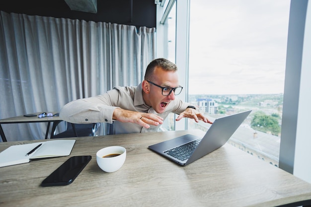 Emotional young manager at work desk with laptop on face glasses A cheerful businessman is working in a modern office with large windows