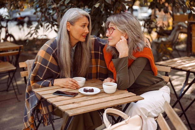 Empathic grey haired asian woman hugs friend to cheer up at table in street cafe