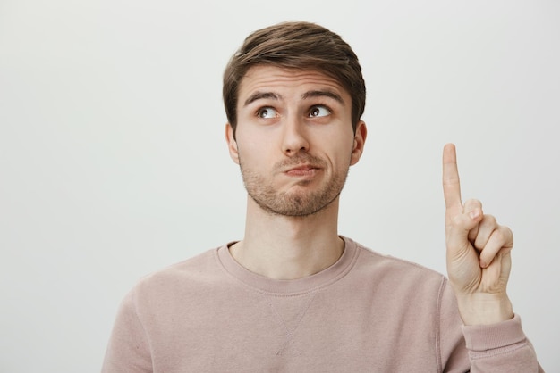 Employee has suggestion Portrait of handsome european male student raising index finger and looking up with puckered lips confessing that it was his fault being embarrassed over gray background