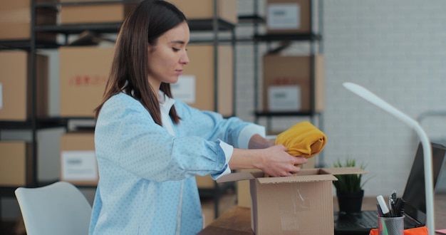 An employee packs a yellow jumper in a room with shelves full of parcels The woman owner of an online store works on a computer at his desk in a warehouse