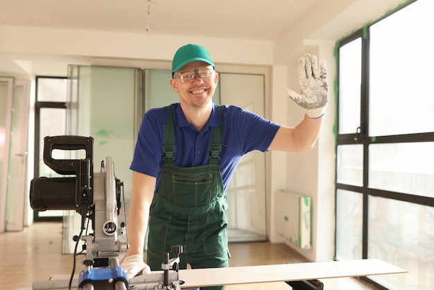 Employee waves hand standing by sawing machine in renovated room happy worker with equipment