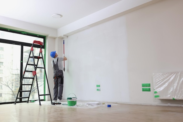 Employee with helmet paints wall with roller brush in spacious premise builder renovates room