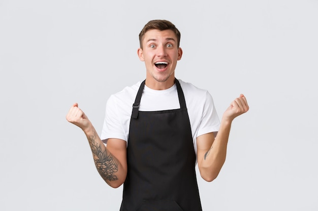 Employees, grocery stores and coffee shop concept. Happy and relieved smiling handsome barista, waiter in black apron, fist pump in rejoice, triumphing victory or great positive news