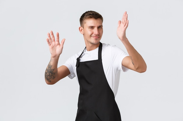 Employees, grocery stores and coffee shop concept. Pleased and happy handsome cheeky barista, guy in black apron, working in cafe as waiter, dancing from joy and clap hands, white background