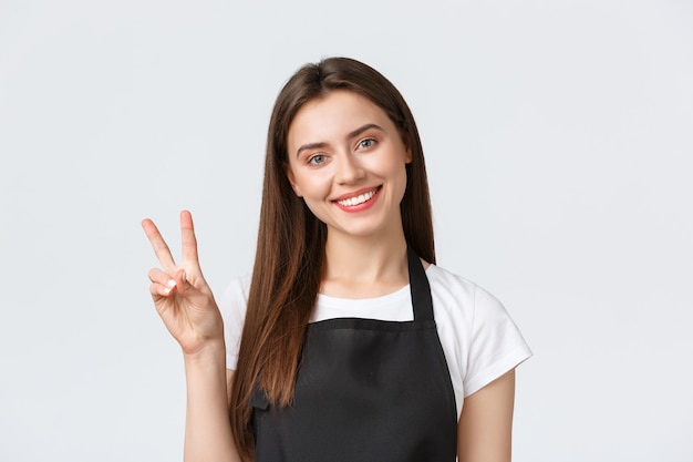 Employees, job employment, small business and coffee shop concept. Close-up of friendly-looking smiling female barista, cafe worker in black apron greeting guests and show peace sign.