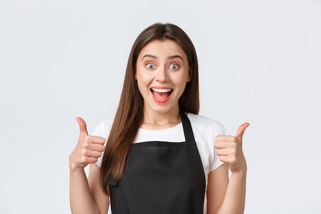 Employees, job employment, small business and coffee shop concept. Satisfied cute female saleswoman in black apron showing thumbs-up, guarantee provide best service, recommend buy in this store.