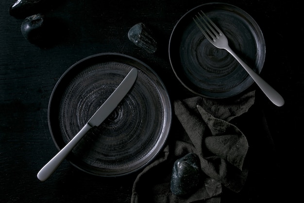 Empty black ceramic plates with cutlery and black stones around on textile napkin over black wooden background. Flat lay, space.