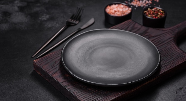 An empty black plate on a dark concrete background with spices herbs and cutlery