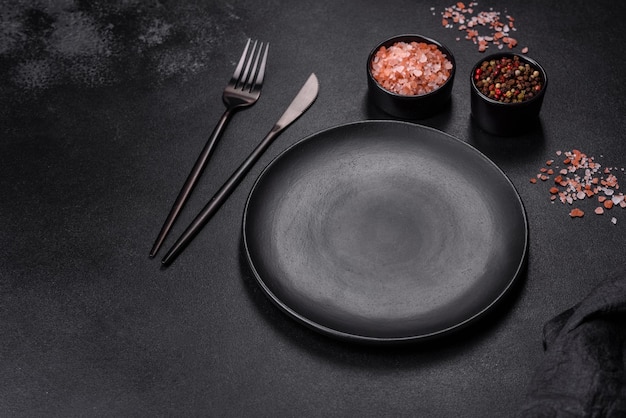 An empty black plate on a dark concrete background with spices herbs and cutlery