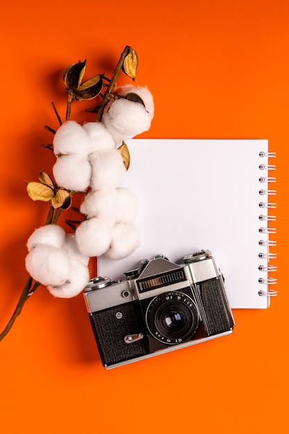 A empty blank notebook on an orange desk, cotton branch, and a vintage camera