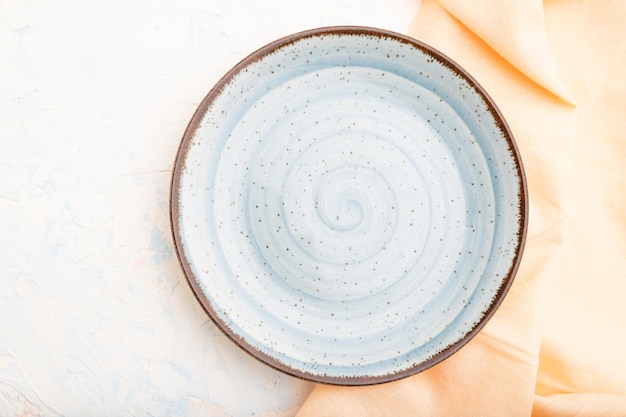 Empty blue ceramic plate on white concrete background and orange linen textile. Top view, close up, flat lay.