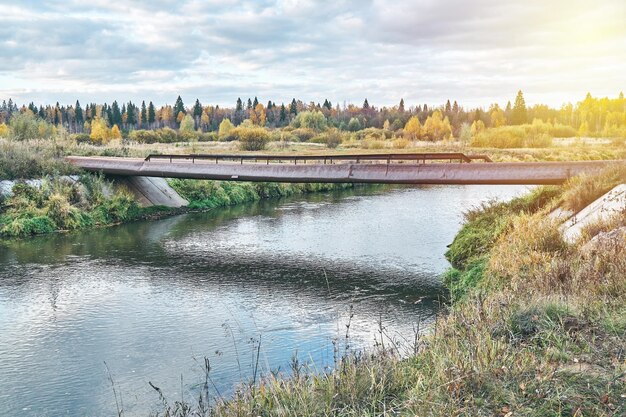 Empty bridge made of old metal pipes with small railing over tranquil river at rural site