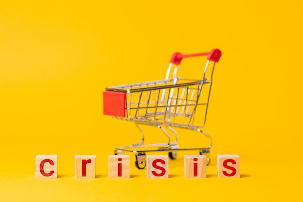 An empty cart and wooden blocks, with a red text crisis. Yellow background. The concept of economy, crisis and shopping