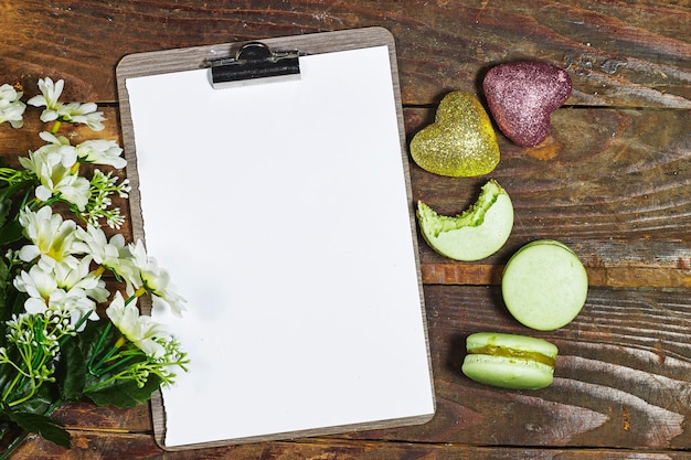 Empty clipboard with daisy flowers and macarons and heart glitters on wood table