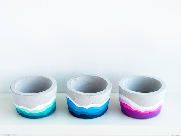 Empty colorful DIY round concrete pots on a white wooden shelf on white wall with copy space. Three unique colorful color painted cement planters.