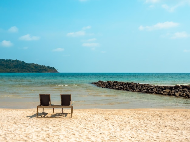 Empty couple sunbed seats on the sandy beach in front of the blue sea background near the rocks at the island with nobody Relaxing two chairs on seascape background on sunny day