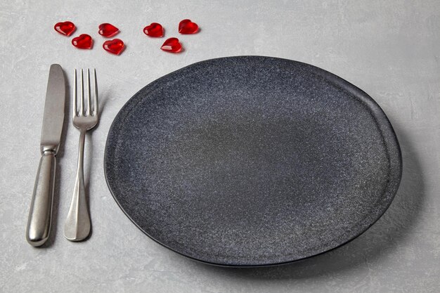 Empty dark plate with cutlery and red caramel hearts on a light concrete table Romantic menu Top view