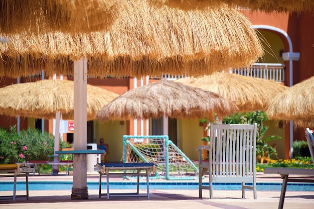 Empty deck chairs under straw shade umbrellas on swimming pool side in tropical resort Summer vacations and getaway concept