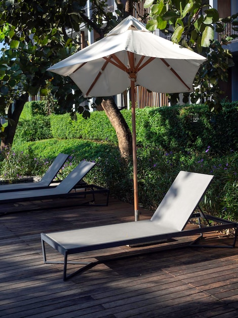 Empty fabric sunbeds and beach umbrella on the wood deck floor near the swimming pool and outdoor garden near the building with nobody, vertical style. Summer background and recreation concept.