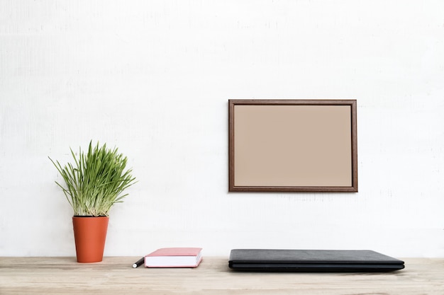 Empty frame on white wall. Closed laptop, notebook and pen. Green houseplant. Home workplace.