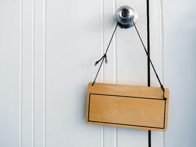 Empty frame with blank space for text on vintage wooden signboard with rope hanging on old door knob on white wood door background with copy space closeup