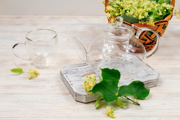 Empty glass teapot and two cups for tea on a wooden table against a background of lime leaves and flowers Healing tea