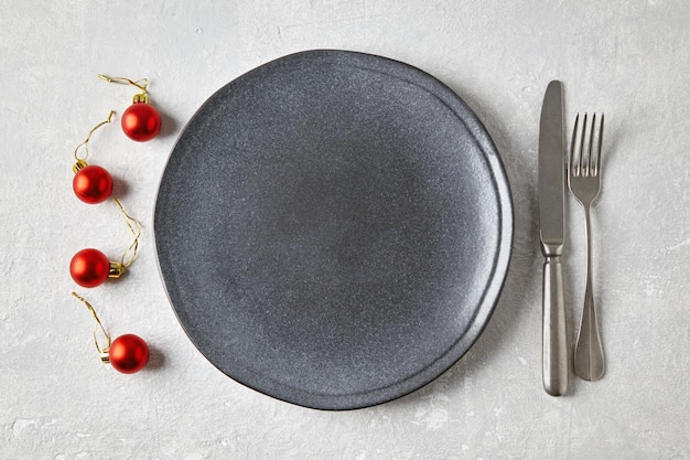 An empty gray ceramic plate with cutlery surrounded by red Christmas balls on a light concrete table Mockup for displaying food