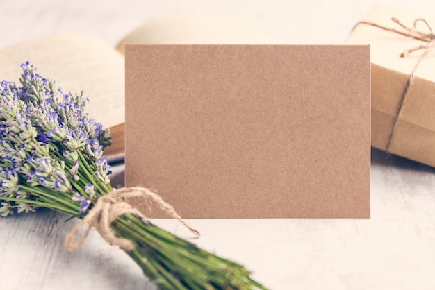 Empty greeting kraft card in front of a lavender bouquet, wrapped gift and old book over a white wood background. 