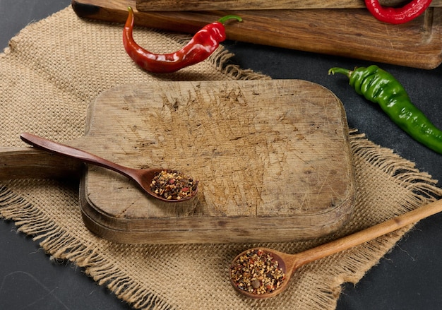 Empty kitchen brown wooden cutting board on black table, next to spoons with spice and fresh chili, black background