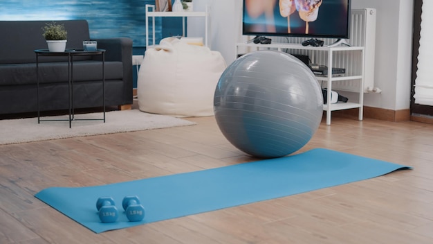 Empty living room with equipment used for physical exercise. Nobody in space with yoga mat, dumbbells and stretching fitness ball to workout and practice sport. Gymnastics and wellness