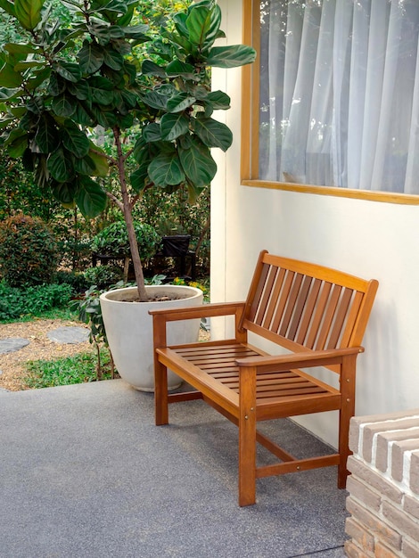 Empty minimal style cozy wooden bench with arms decorated on a white wall near the big concrete pot outside the building near a window and the green garden vertical style