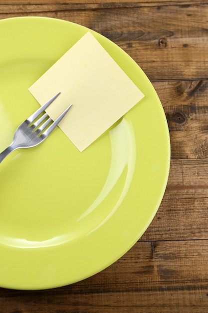 Empty note paper attached to fork on plate on color wooden background