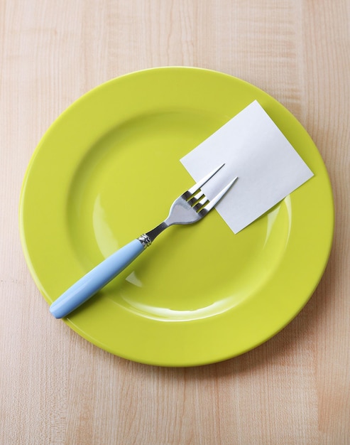 Empty note paper attached to fork on plate on color wooden background