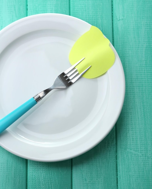 Empty note paper attached to fork on plate on color wooden background