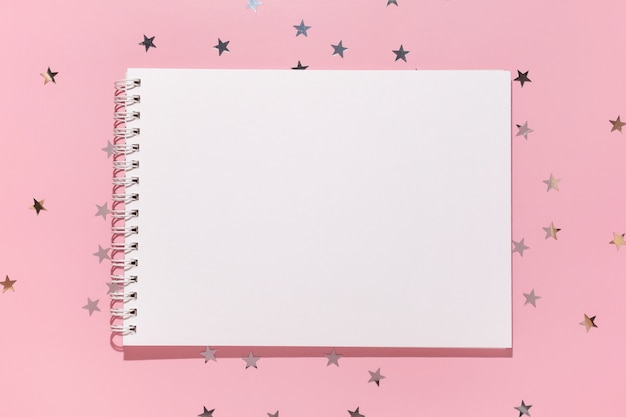 Empty notebook and confetti stars pours on a pastel pink background, top view. Flat lay. Holiday, greeting or good news concept. Mockup template. top view. Blank notepad