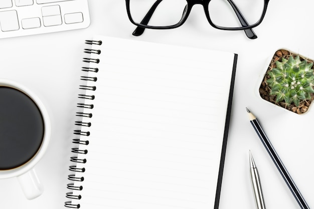 Empty notebook page is on top of white office desk table with supplies