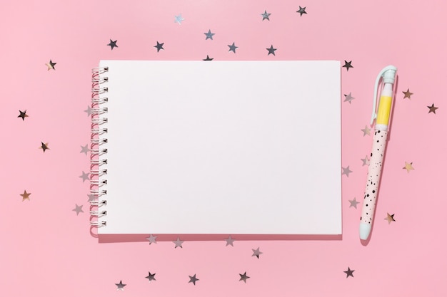 Empty notebook with pen and confetti stars pours on a pastel pink background, top view. Flat lay. Holiday, greeting or good news concept. Mockup template. top view. Blank notepad