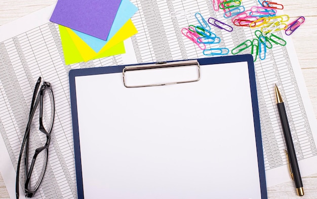 An empty notebook with a pen reports bright stickers paper clips and glasses lies on a white office table Top view with copy space flat lay