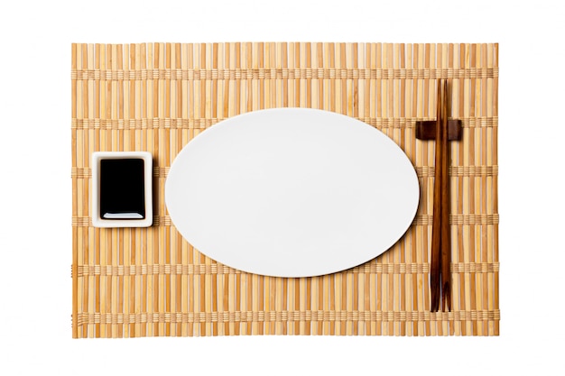 Empty oval white plate with chopsticks for sushi and soy sauce on yellow bamboo mat . 